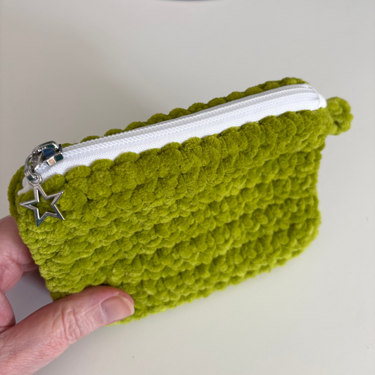 Green crocheted pouch with a star zipper pull held by a hand on a white surface.